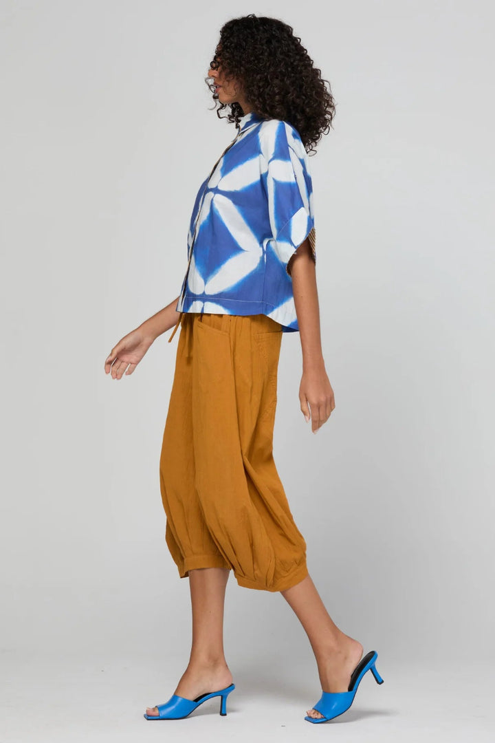 A woman modeling a blue and white geometric patterned short-sleeve top paired with mustard yellow cropped pants and bright blue mule heels, photographed against a light gray studio background.