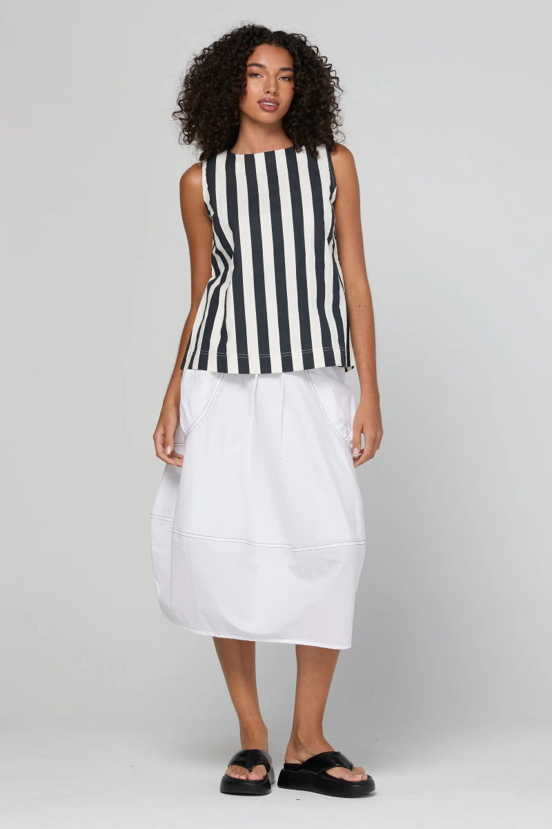 A model stands against a light gray studio background wearing a sleeveless black and white vertically striped top paired with a white midi skirt and black slide sandals.