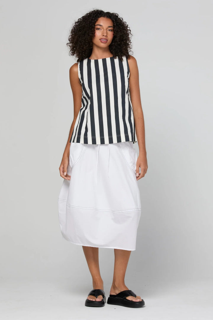 A model stands against a light gray studio background wearing a sleeveless black and white vertically striped top paired with a white midi skirt and black slide sandals.