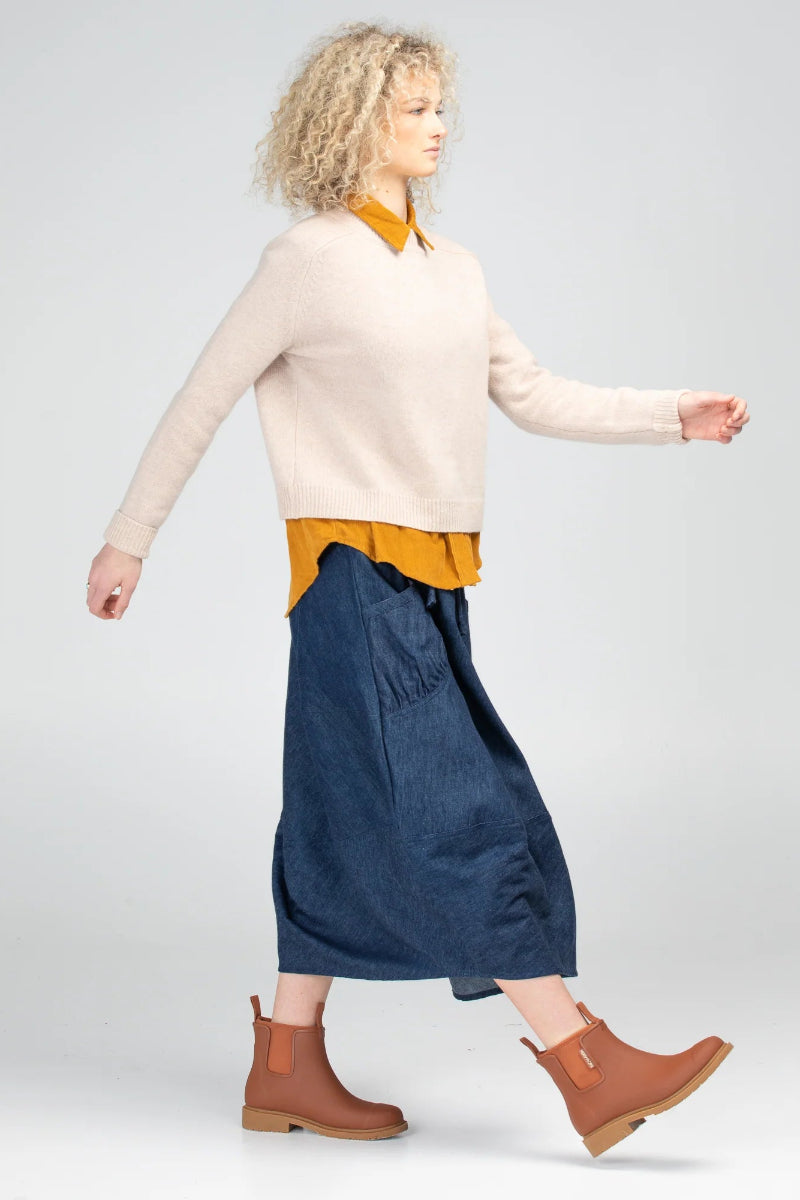 Woman wearing a beige sweater, yellow shirt, blue skirt, and brown boots on a light gray background