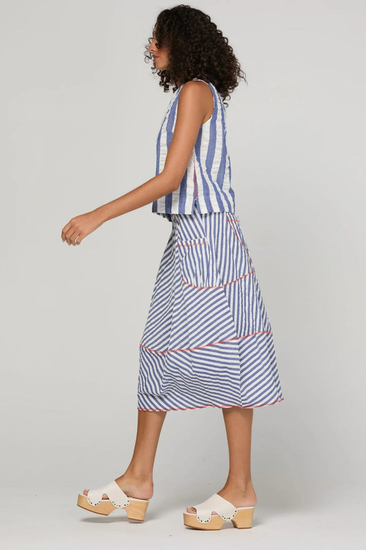 A woman wearing a sleeveless blue and white striped top and matching striped midi skirt with thin red accent lines, paired with white slide sandals, captured from a side profile against a light gray studio background.
