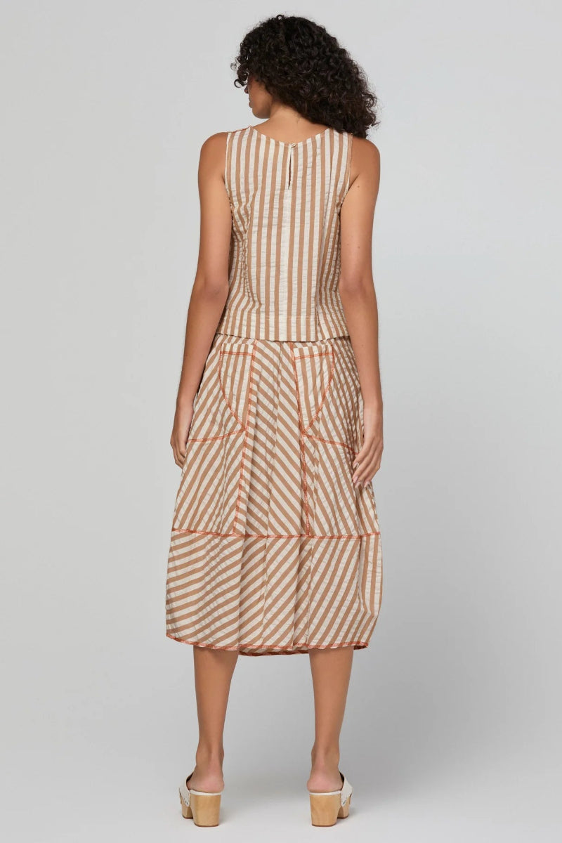 A woman viewed from the back wearing a sleeveless midi dress with vertical beige and rust-orange stripes, paired with beige block-heel mules, against a light gray studio background.