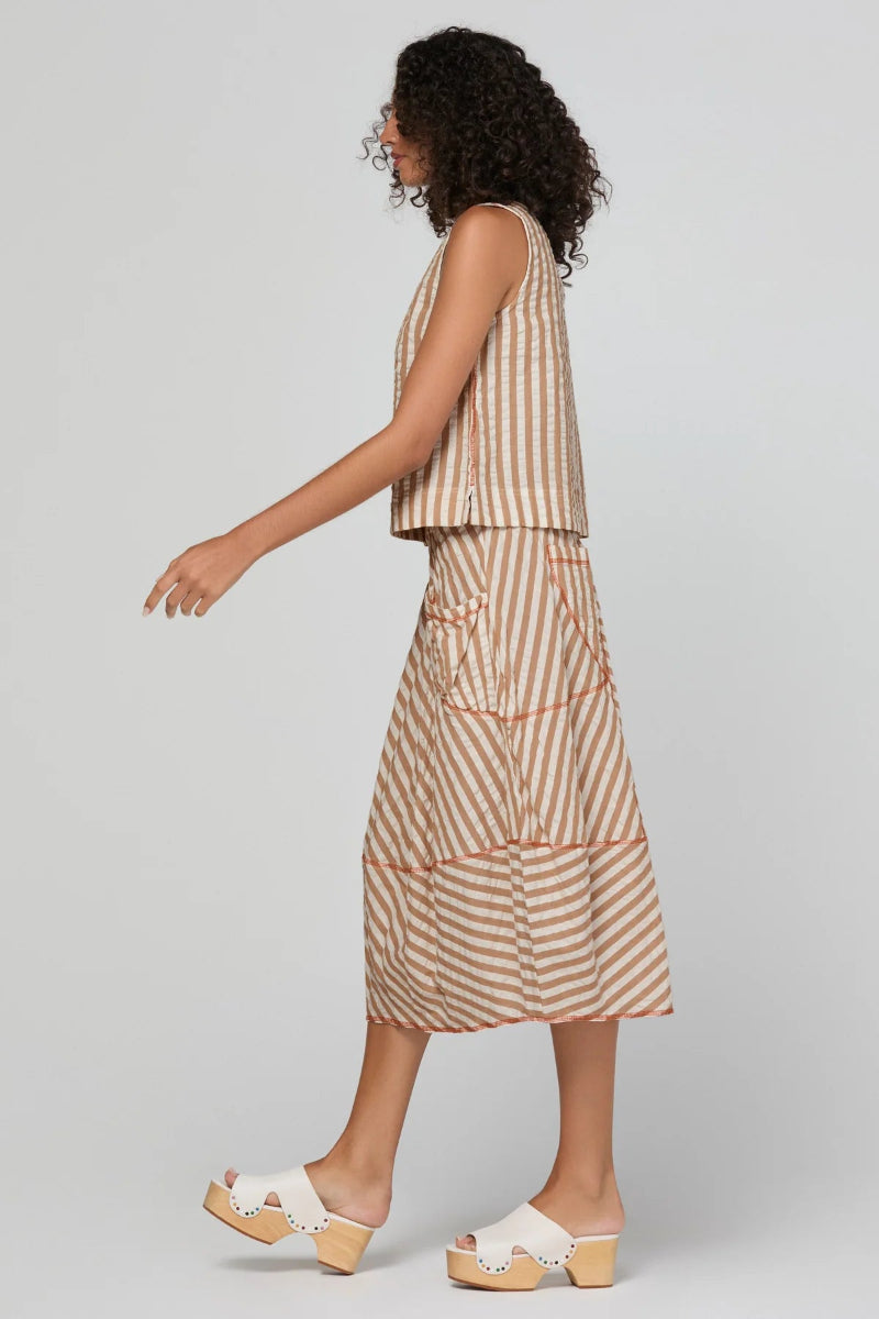 A model with curly dark hair wearing a sleeveless beige and white striped crop top and matching striped wide-leg skirt with a beige drawstring waist, photographed against a light gray studio background.