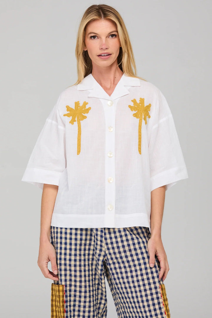 Woman wearing a white shirt with palm tree designs and checkered pants on a gray background