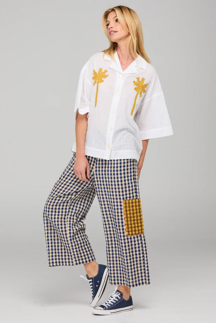 Woman wearing a white oversized shirt with palm tree designs and checkered pants on a gray background