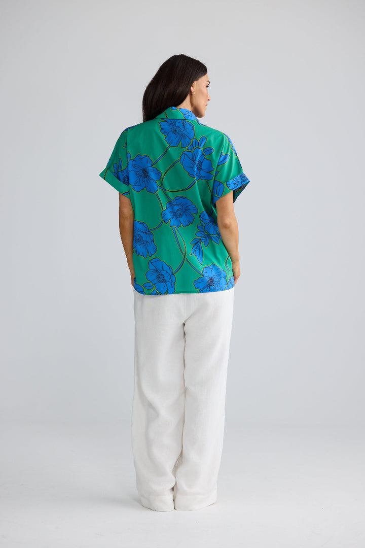 A woman is shown from the back wearing a short-sleeve button-up shirt with a green base and a blue floral pattern, paired with white trousers, against a light gray studio background.