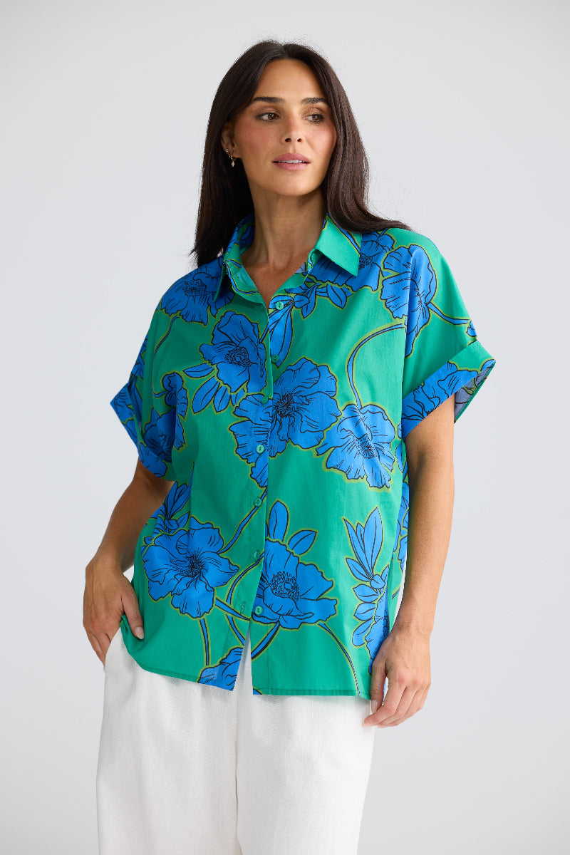 A woman modeling a short-sleeve button-up shirt with a vibrant green base and a bold blue floral pattern outlined in gold, paired with white pants against a light gray studio background.