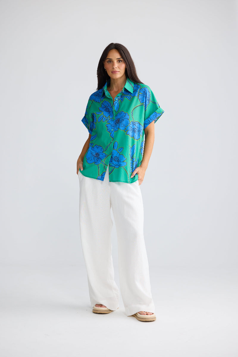 A woman modeling a short-sleeve button-up shirt with a green and blue floral pattern, paired with white wide-leg pants and beige sandals, against a light gray studio background.