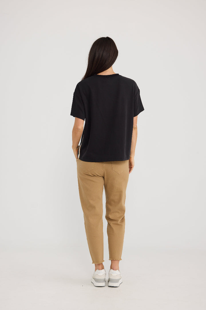 A woman viewed from behind wearing a black short-sleeve t-shirt, tan pants, and white sneakers against a plain light gray studio background.