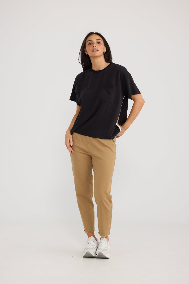A woman modeling a black short-sleeve crewneck t-shirt and tan slim-fit pants with white sneakers, standing against a light gray studio background.