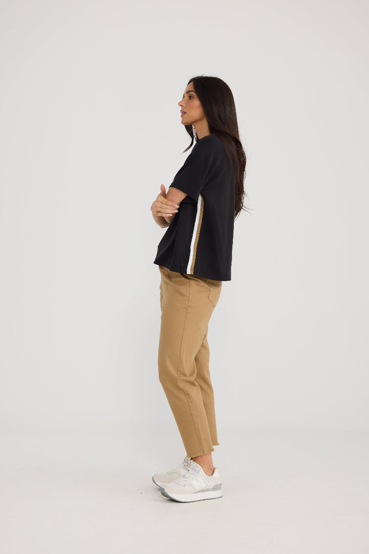 A woman stands in profile wearing a black short-sleeve crop top with a white and gold stripe detail on the sleeve, tan cropped pants, and white sneakers, against a plain light gray studio background.