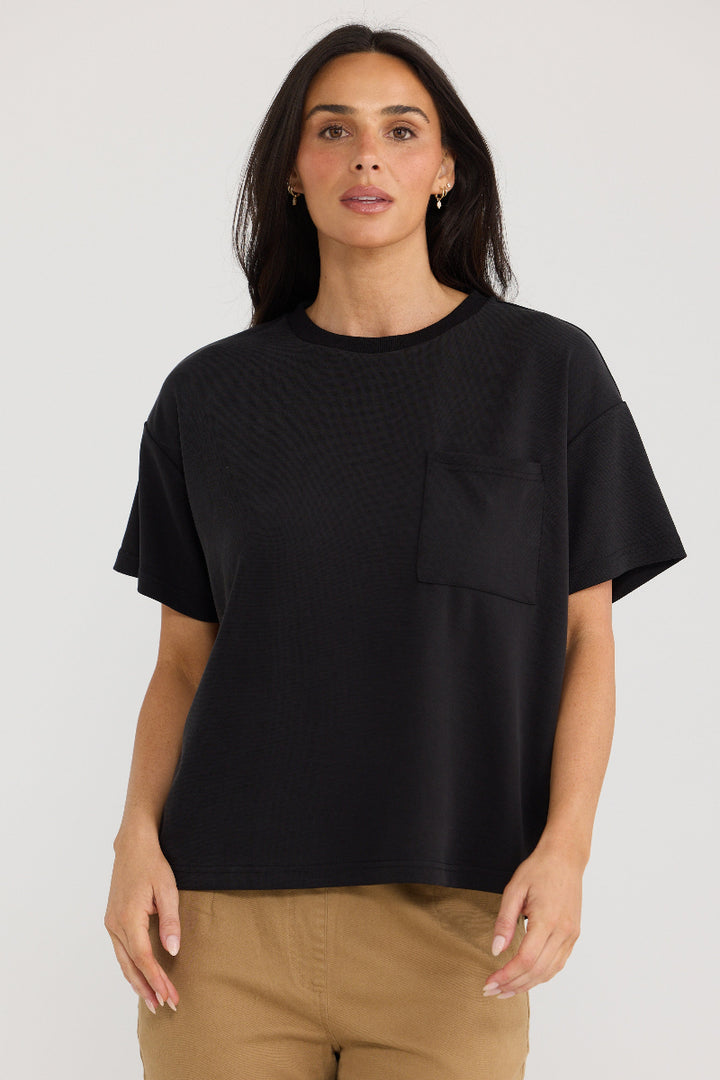 A woman wearing a black short-sleeve crewneck t-shirt with a chest pocket, paired with tan pants, against a light gray studio background.
