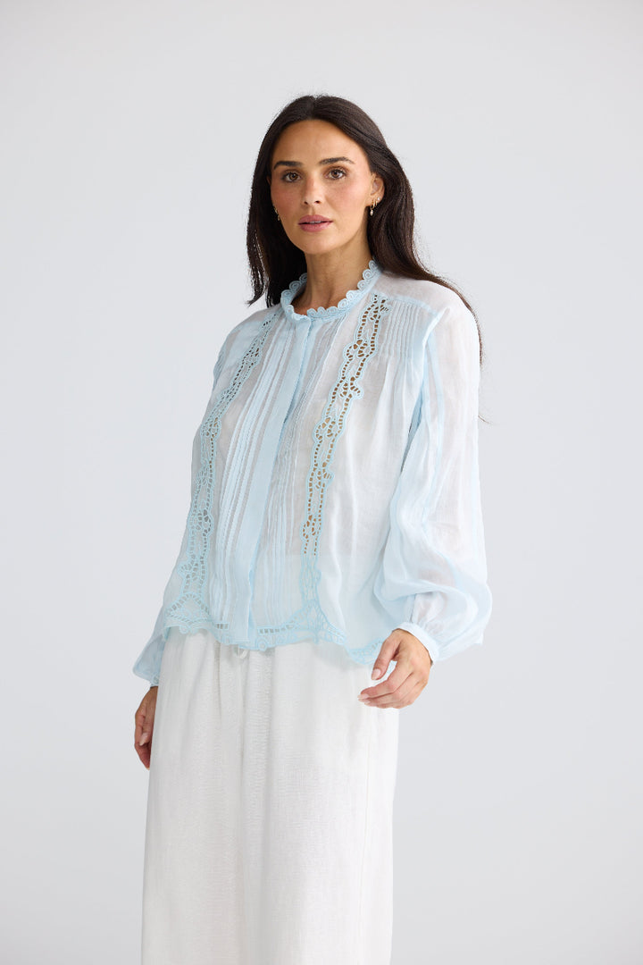 A woman modeling a light blue sheer long-sleeve blouse with vertical embroidered detailing, paired with white wide-leg pants, against a plain light gray studio background.