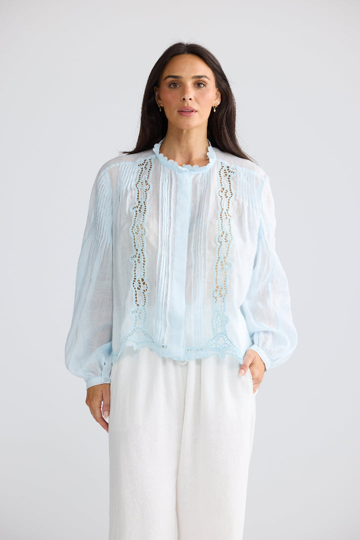 A woman modeling a light blue sheer long-sleeve blouse with vertical embroidered detailing, paired with white wide-leg pants, against a plain light gray studio background.