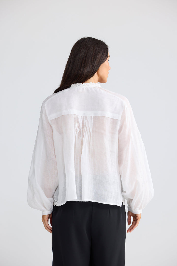 A woman viewed from the back wearing a white sheer long-sleeve blouse with gathered pleating at the back and black pants, against a light gray studio background.