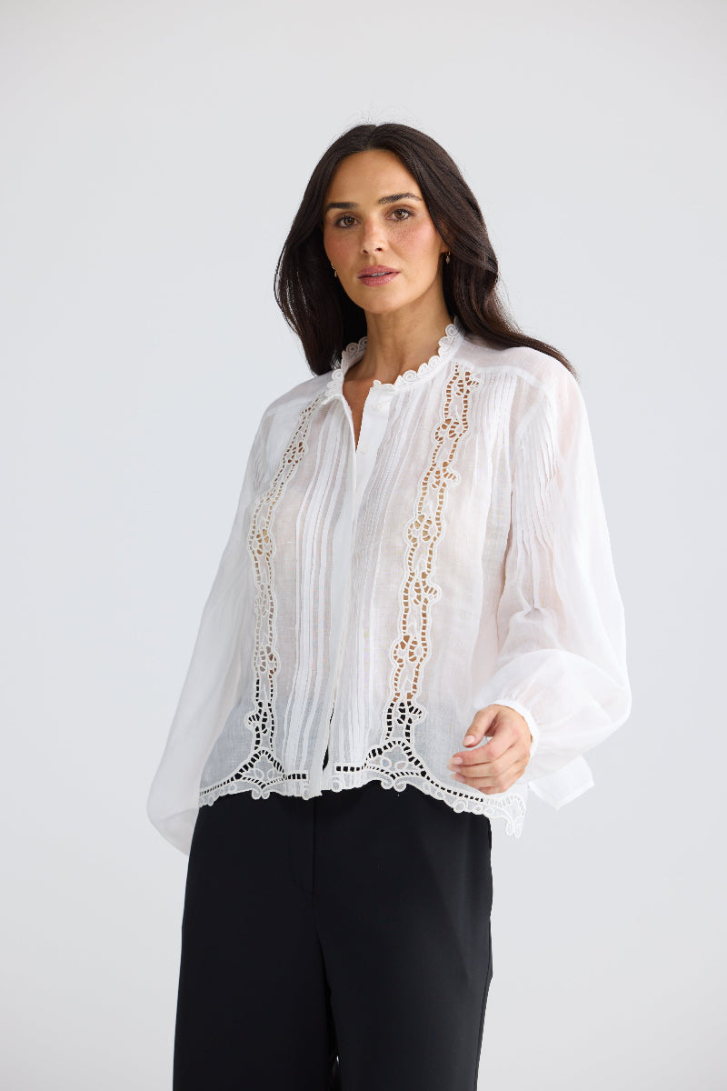 A woman modeling a white long-sleeve blouse with delicate gold embroidery and lace detailing, paired with black trousers, against a light gray studio background.