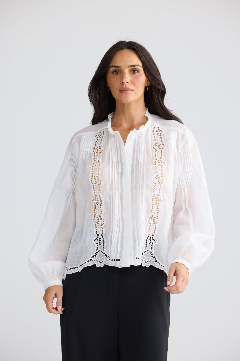 A woman modeling a white long-sleeve blouse with vertical lace applique panels down the front, paired with black trousers, against a light gray studio background.