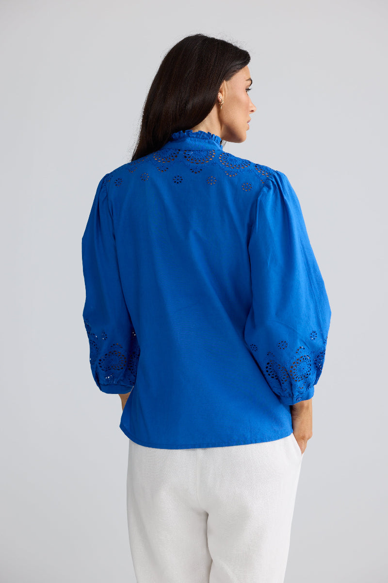 A woman shown from the back wearing a bright blue blouse with eyelet embroidery on the shoulders and sleeves, paired with white pants, against a light gray studio background.