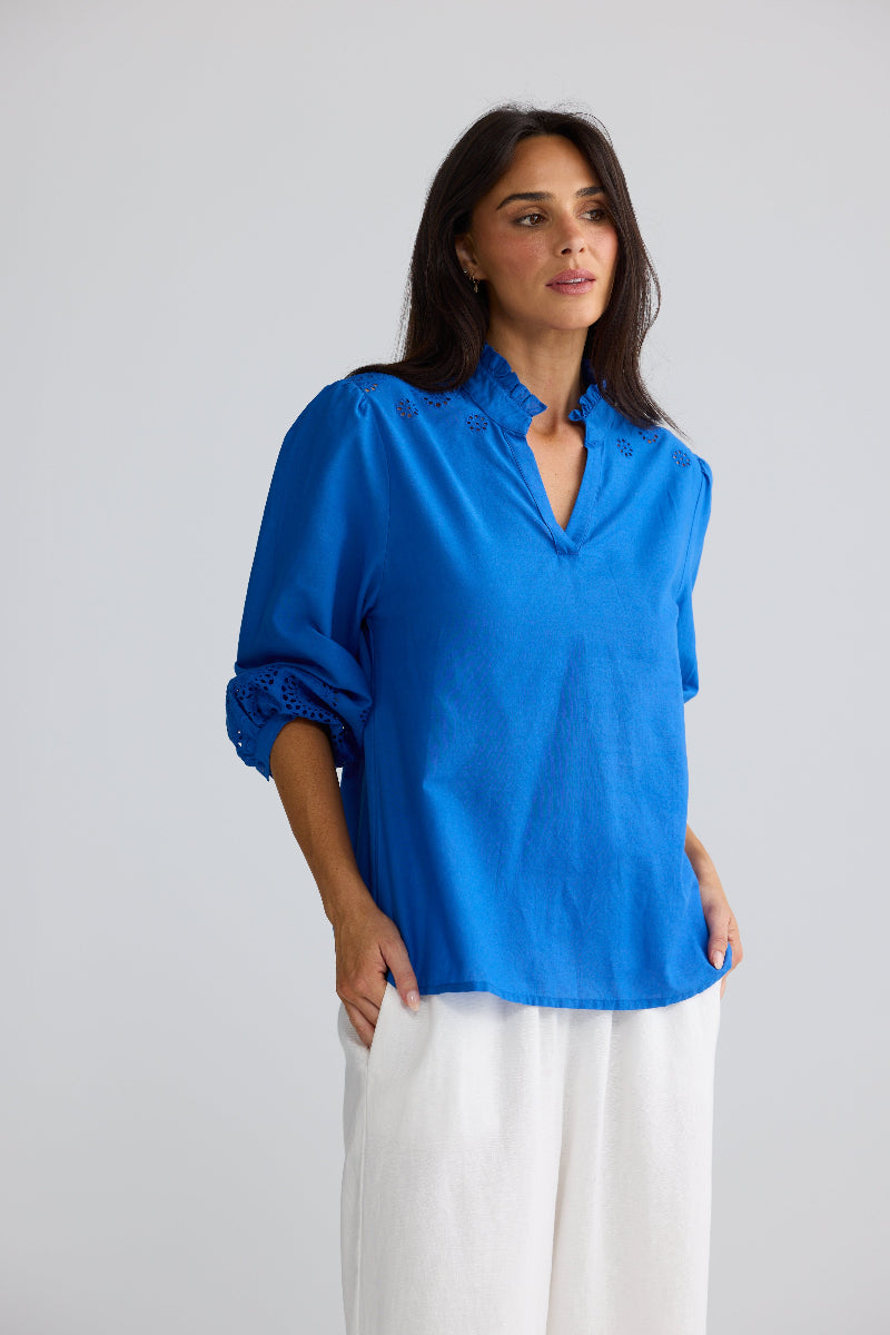 A woman modeling a bright blue linen blouse with rolled sleeves and white wide-leg trousers, standing against a light gray studio background.