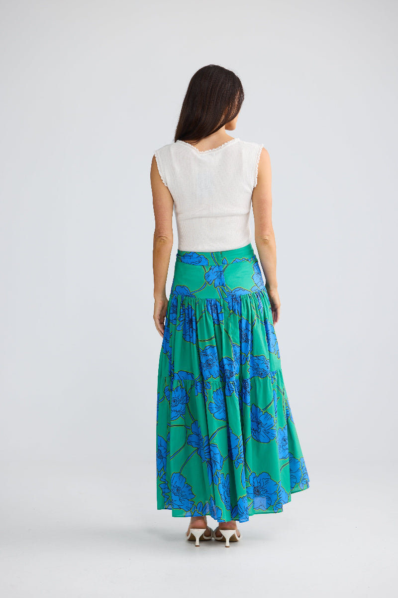 A woman shown from the back wearing a sleeveless white top and a long green skirt with a blue floral print, photographed against a light gray studio background.