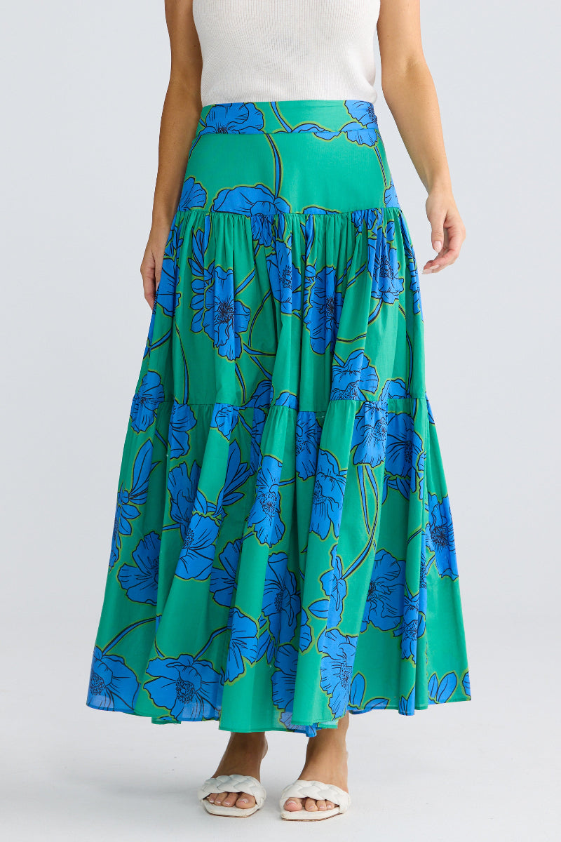 A model wearing a high-waisted, tiered maxi skirt with a vibrant green and blue floral pattern, paired with a white top and white slide sandals, against a light gray studio background.