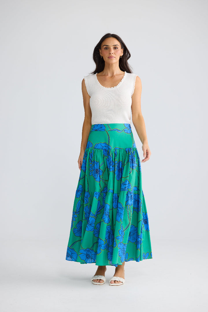A woman stands against a light gray studio background wearing a sleeveless white top and a long pleated skirt with a green and blue floral pattern.