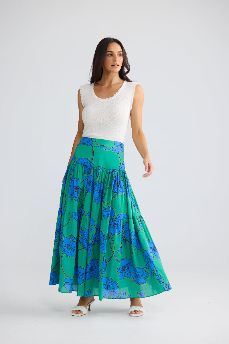 A woman modeling a long, pleated skirt with a blue floral pattern on a green base, paired with a white sleeveless top, against a light gray studio background.