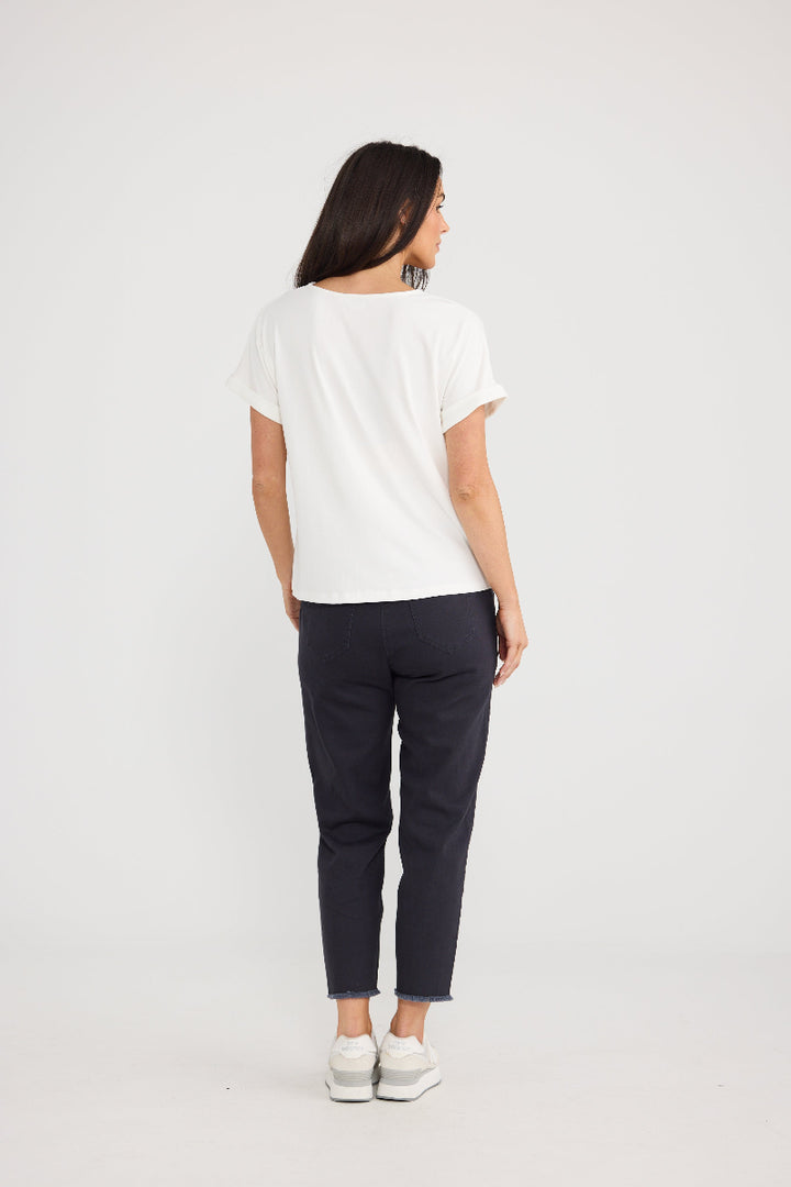 A woman is shown from behind wearing a white short-sleeve t-shirt, dark navy cropped trousers, and white sneakers, standing against a plain light gray studio background.