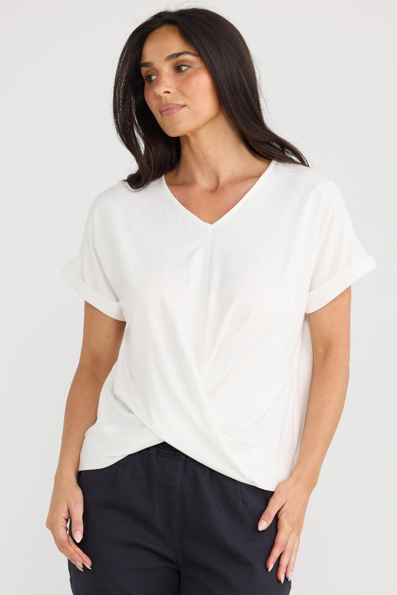 A woman wearing a white short-sleeve V-neck top with a twisted front detail, paired with dark pants, against a light gray studio background.