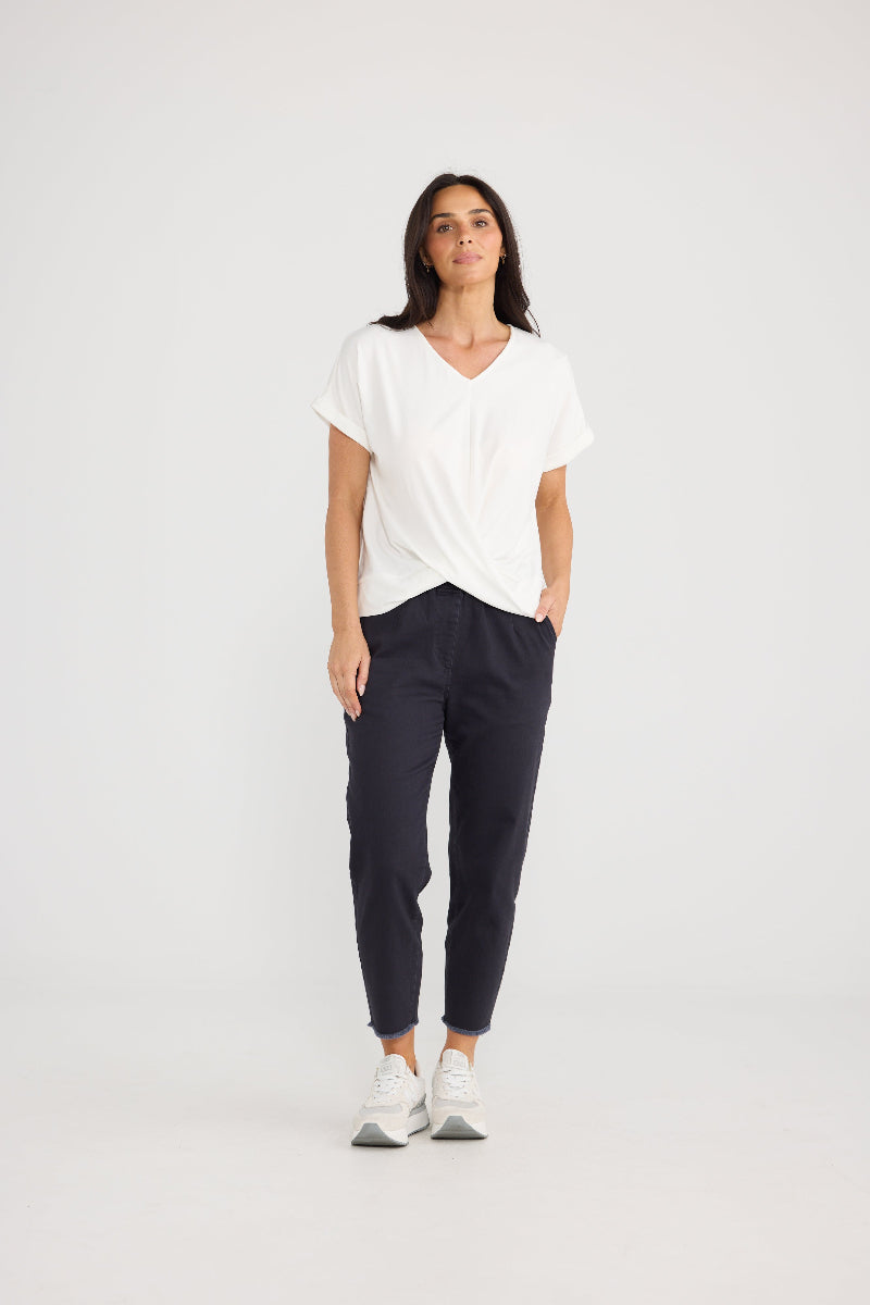 A woman modeling a white short-sleeve V-neck t-shirt and dark navy blue cropped pants, paired with white sneakers, against a plain light gray studio background.