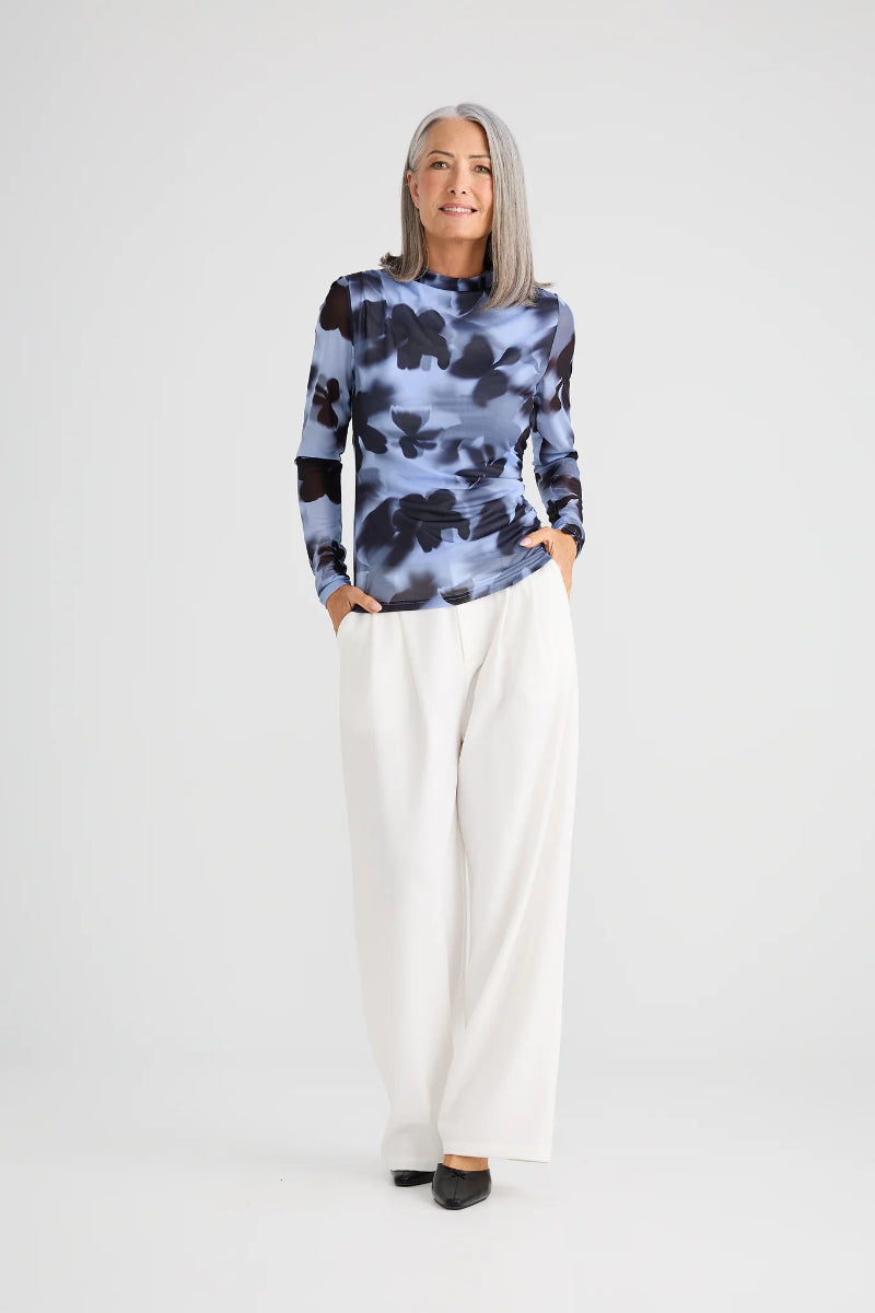 Woman wearing a patterned top and white pants on a plain background