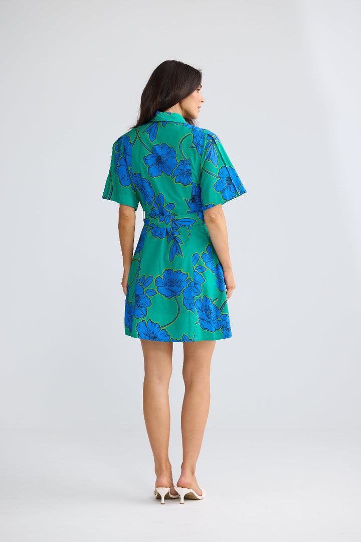 A woman is shown from the back wearing a short-sleeve collared dress with a green base and a blue floral pattern, paired with white heeled sandals, against a light gray studio background.