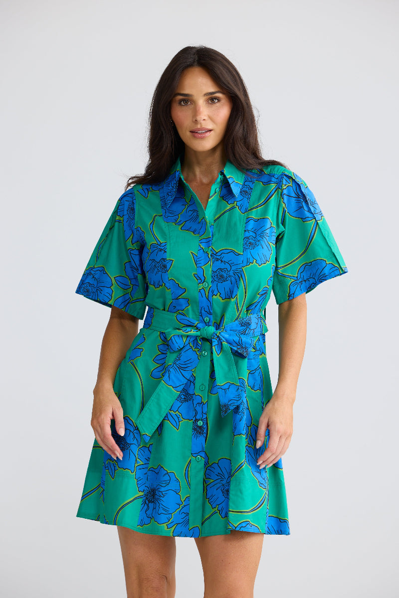 A woman modeling a short-sleeve collared mini dress with a green base and a blue floral pattern, featuring a matching tie belt at the waist, set against a plain light gray studio background.
