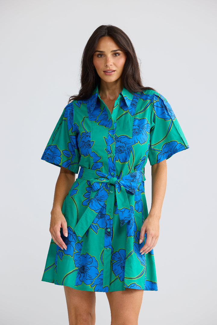 A woman modeling a short-sleeve collared mini dress with a green base and a blue floral pattern, featuring a matching tie belt at the waist, set against a plain light gray studio background.