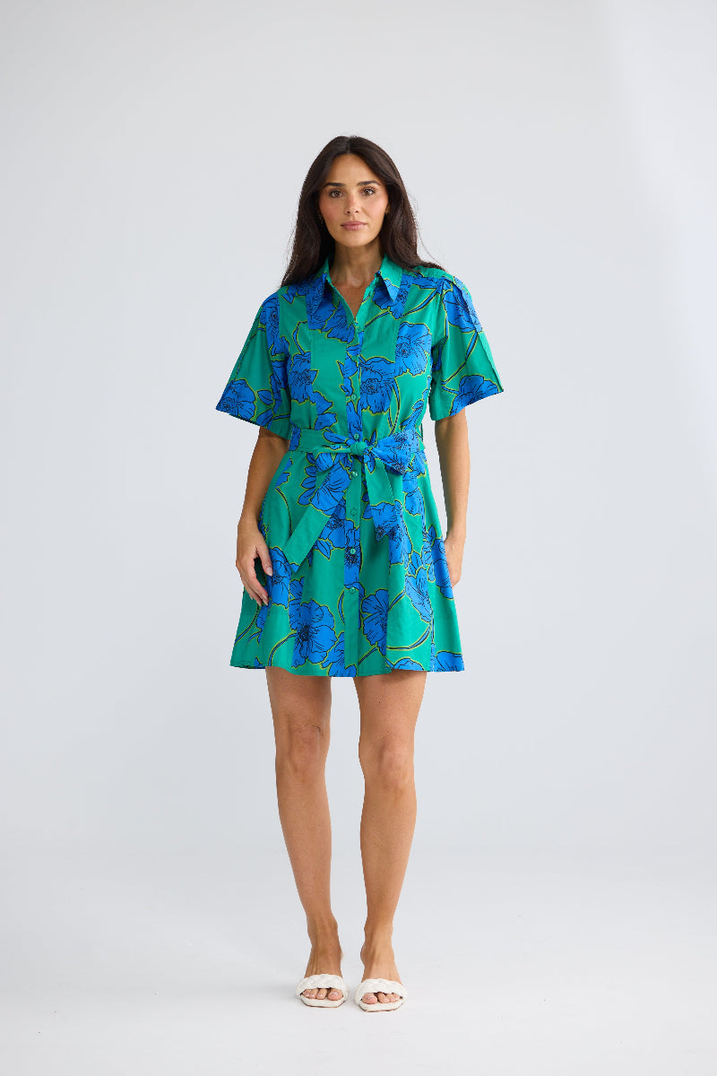 A woman modeling a short-sleeve collared shirt dress with a matching tie waist, featuring a vibrant green and blue floral pattern, set against a plain light gray studio background.