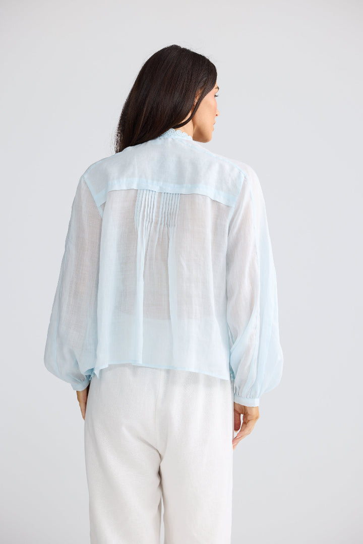 A woman viewed from the back wearing a light blue sheer long-sleeve blouse with gathered pleats at the back and white pants, against a plain light gray studio background.