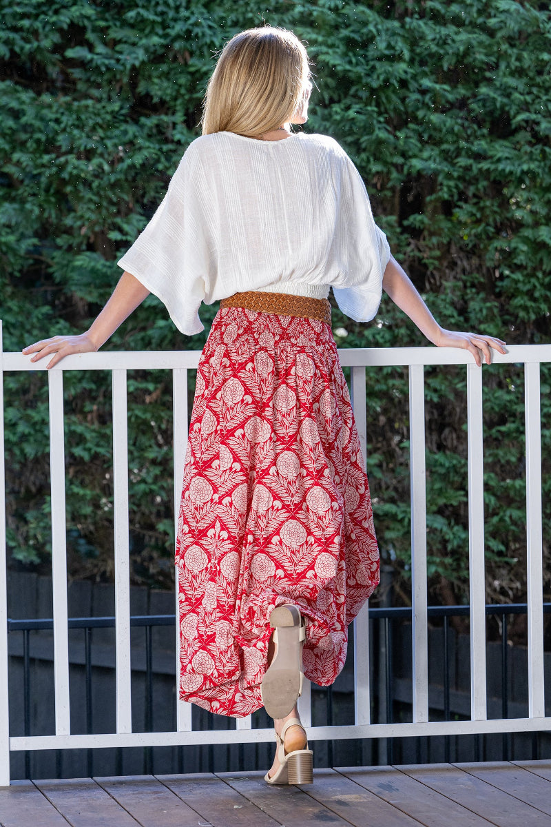 Woman wearing a white blouse and long red, floral patterned skirt standing on a wooden deck with greenery in the background.