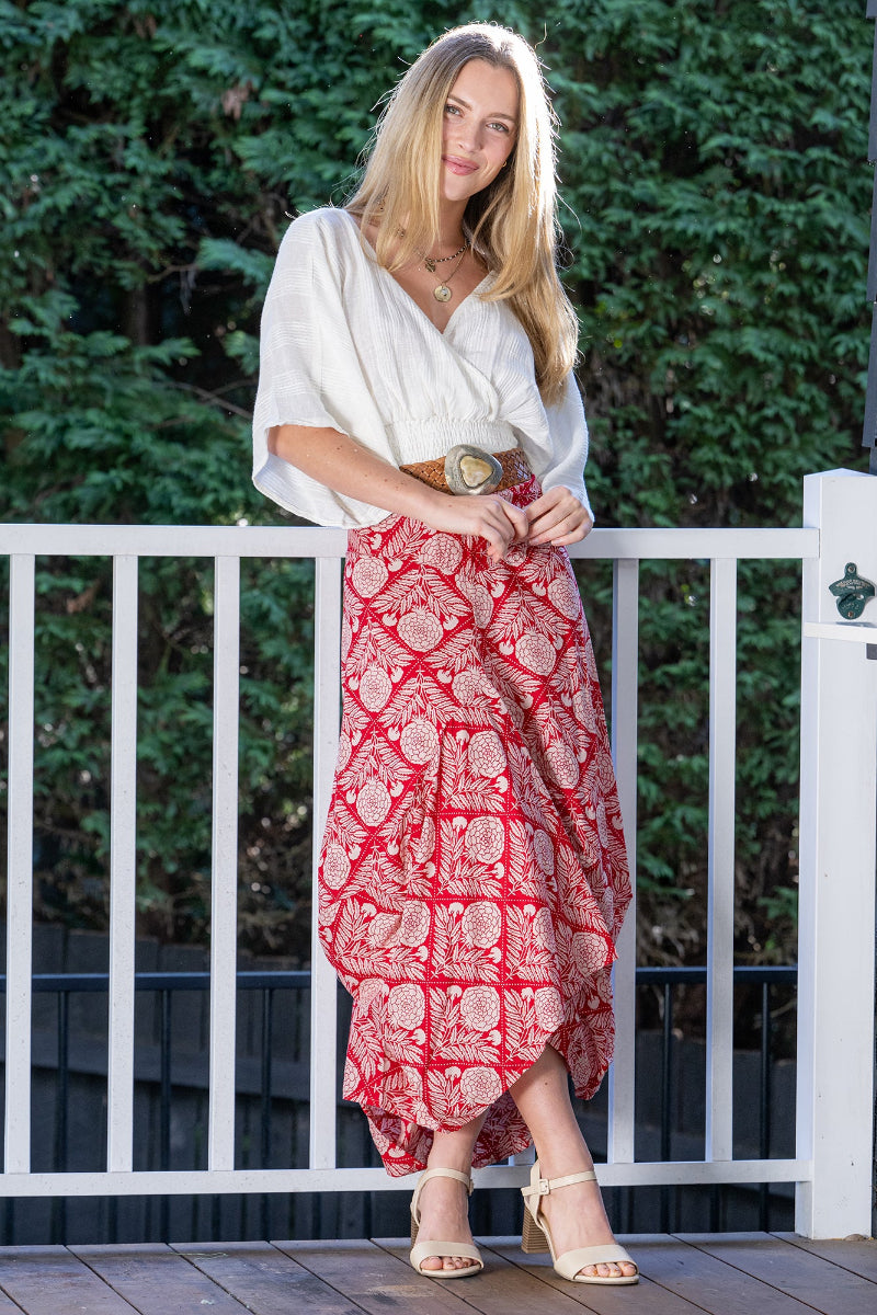 Woman wearing a white blouse and long red, floral patterned skirt standing on a wooden deck with greenery in the background.