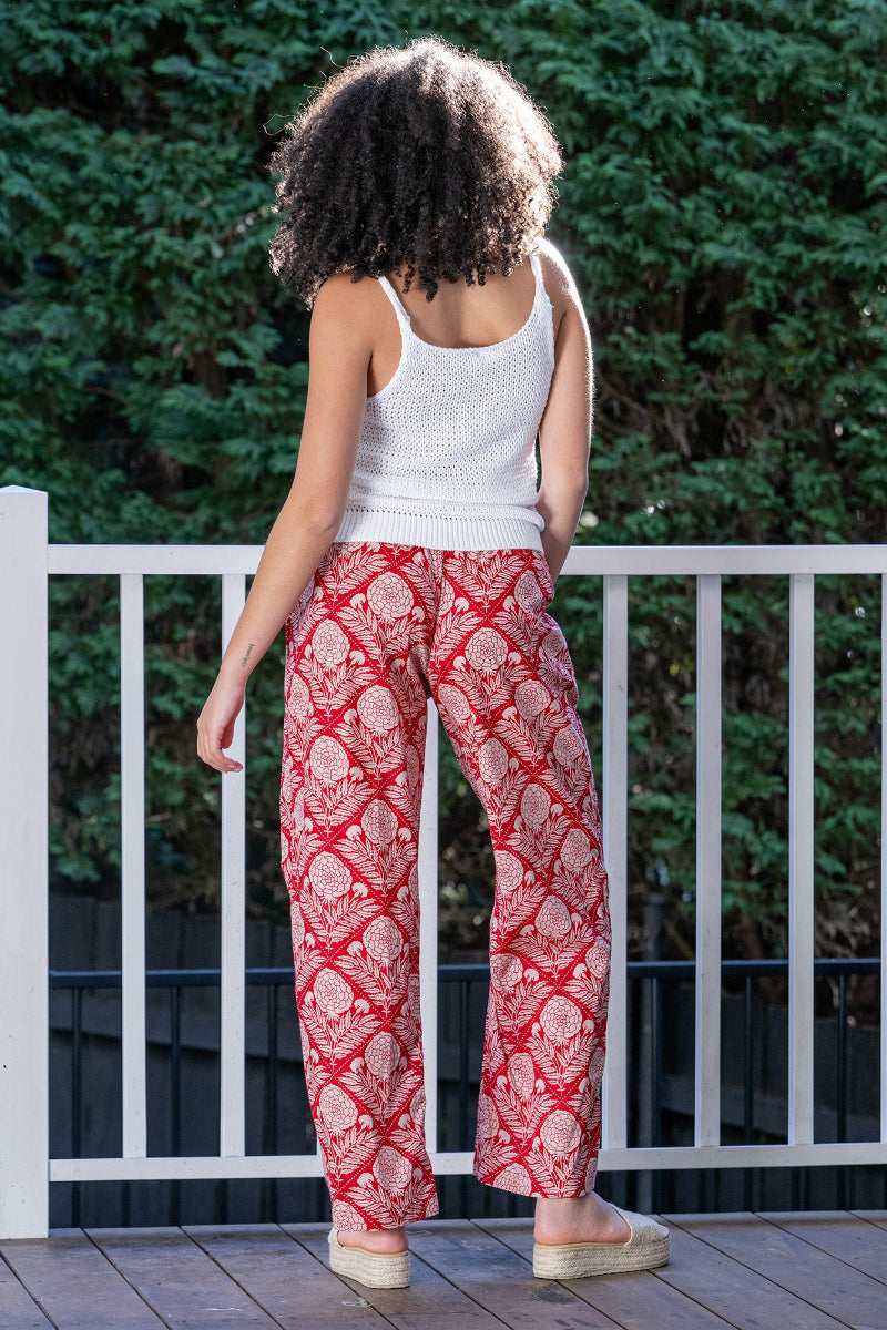 Woman wearing a white sleeveless top and red floral patterned pants standing on a wooden deck with greenery in the background. The pants are full length and flowy, with side pockets and an elastic waistband.