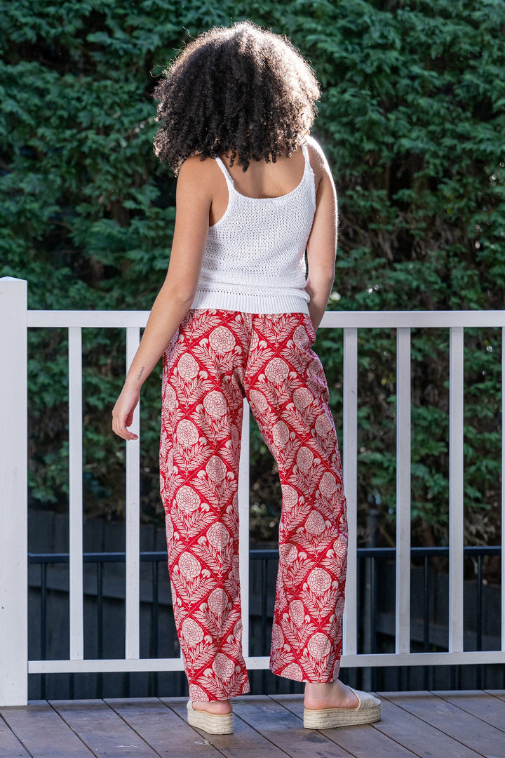 Woman wearing a white sleeveless top and red floral patterned pants standing on a wooden deck with greenery in the background. The pants are full length and flowy, with side pockets and an elastic waistband.