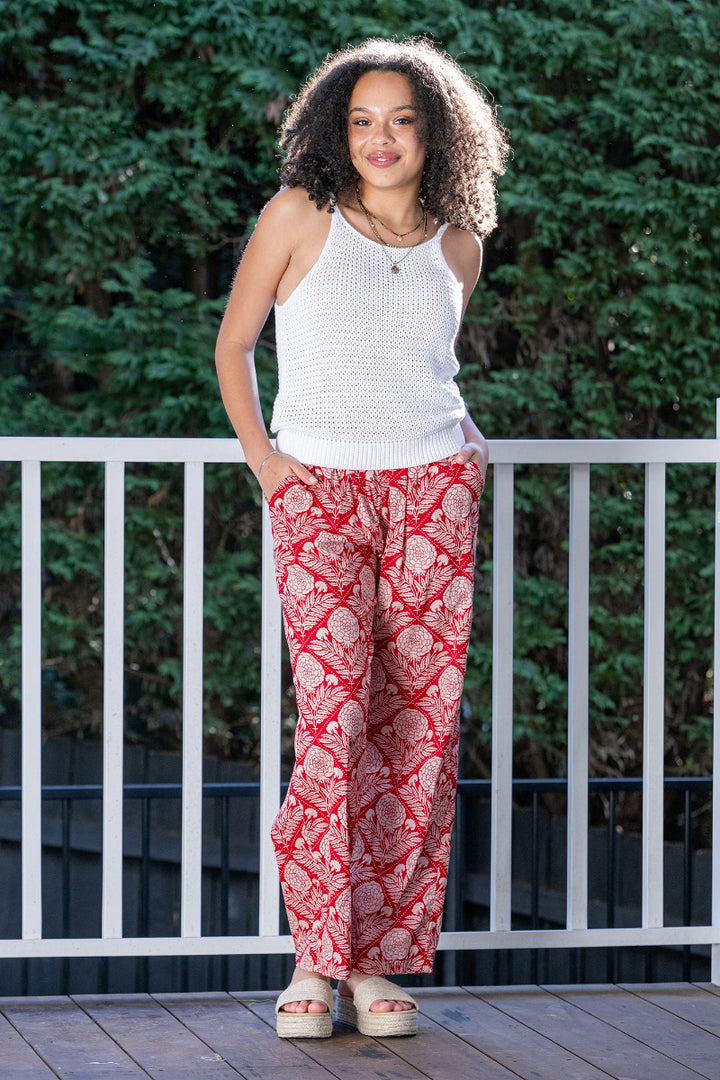 Woman wearing a white sleeveless top and red floral patterned pants standing on a wooden deck with greenery in the background. The pants are full length and flowy, with side pockets and an elastic waistband.