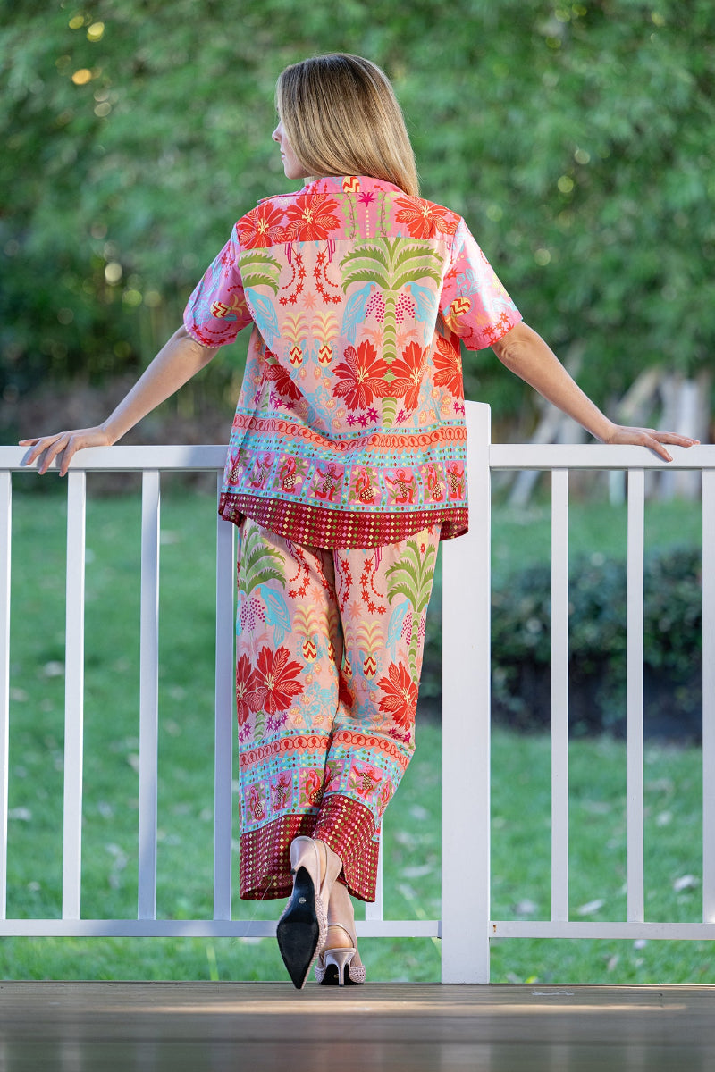 Woman wearing a colorful, floral outfit sitting on a white railing with a green outdoor background. The shirt is collared and features a button up style. The pattern is tropical, with palm trees and hibiscus flowers.