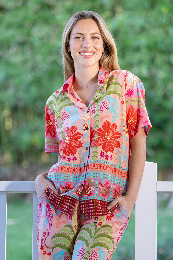 Woman wearing a colorful, floral outfit sitting on a white railing with a green outdoor background. The shirt is collared and features a button up style. The pattern is tropical, with palm trees and hibiscus flowers.