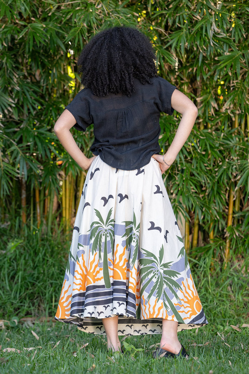Woman wearing a black top and a long skirt with palm tree and sun design, standing in front of green foliage. The skirt has an a-line silhouette.