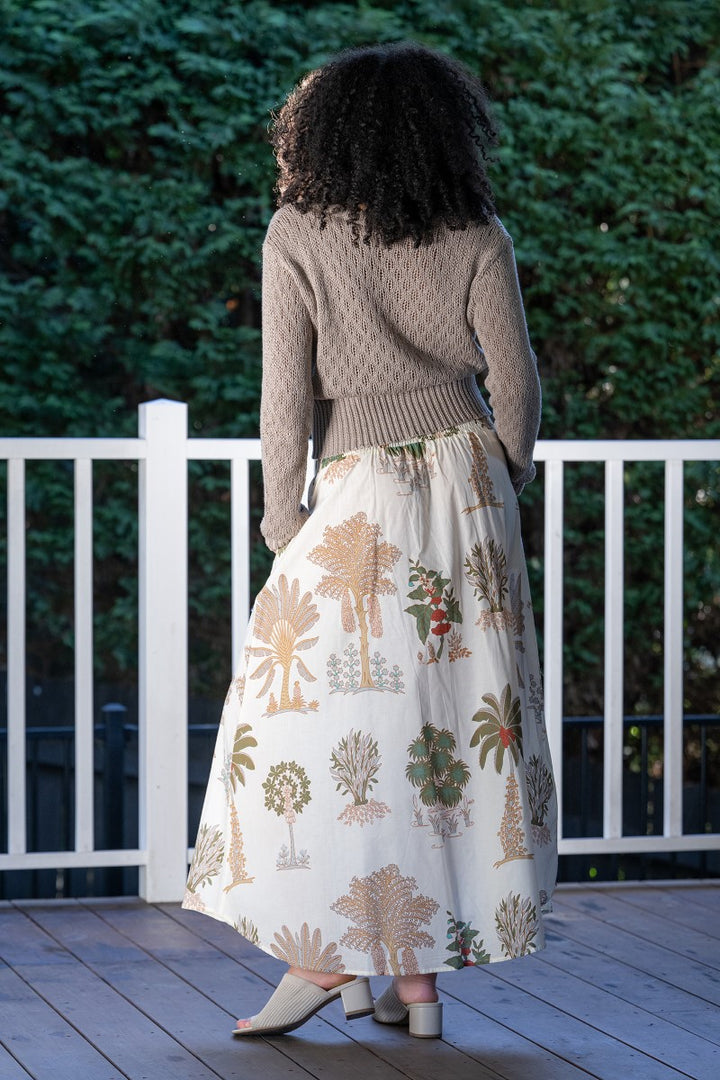 Woman wearing a patterned skirt and sweater on a wooden deck with greenery in the background