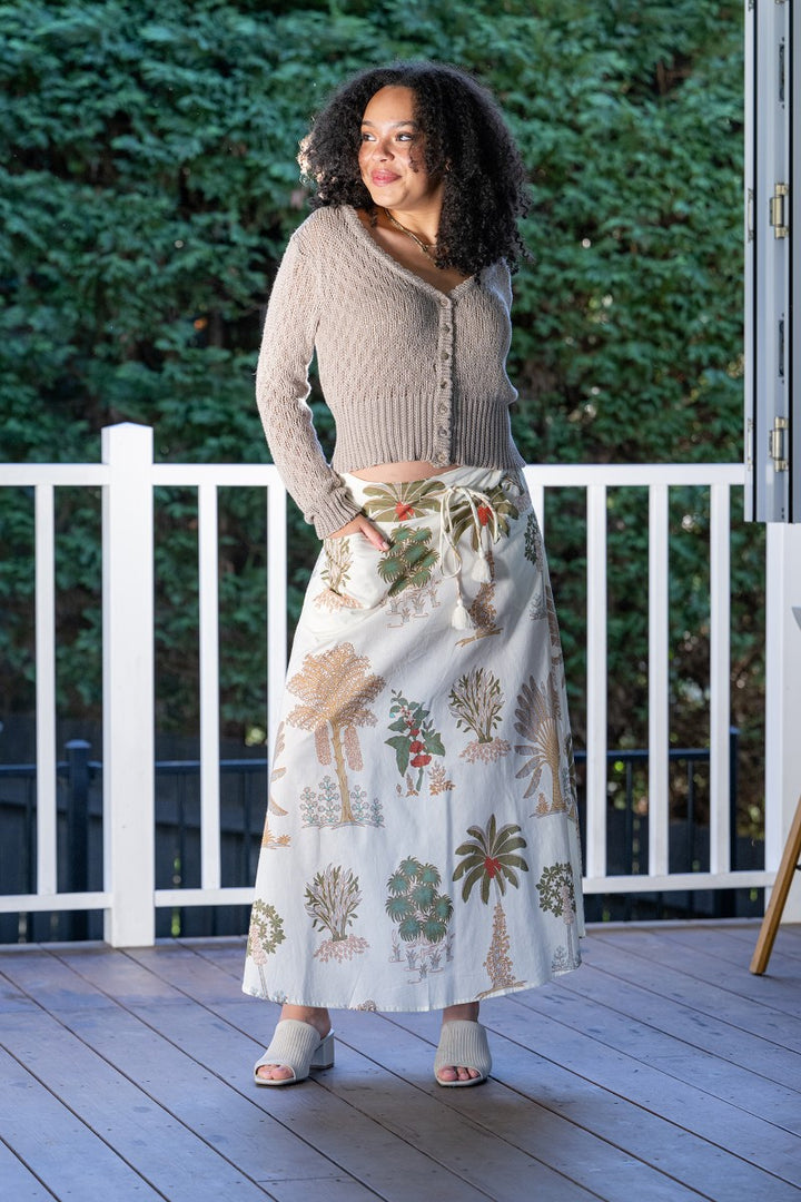 Woman standing on a wooden deck wearing a patterned skirt and beige cardigan.