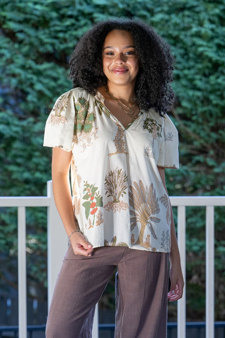 Woman wearing a floral blouse and brown pants standing outdoors with greenery in the background