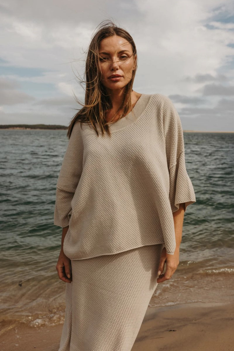 Woman in a beige outfit standing on a beach with ocean and cloudy sky in the background