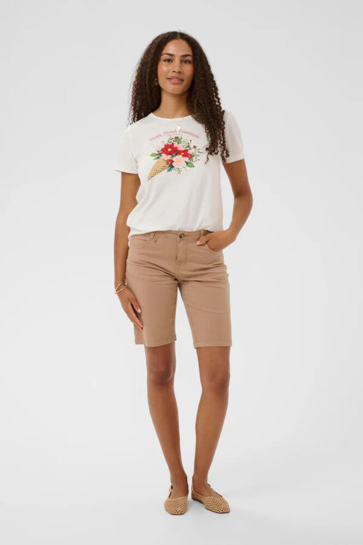 A woman modeling a white short-sleeve graphic T-shirt with a red and green floral/ice cream print, paired with tan shorts and beige flat shoes, against a light gray studio background.