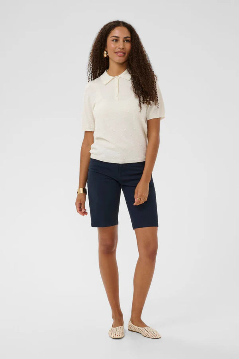 A woman modeling a short-sleeve white polo shirt and navy blue shorts against a light gray studio background.